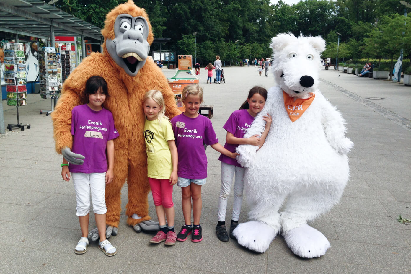 Vier Kinder die mit einem Eisbär-Maskottchen und einem Affen-Maskottchen Händchen haltend und umarmend vor einem Kiosk stehen.